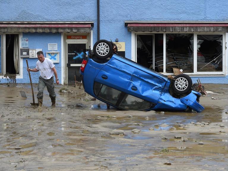 Ein Einwohner des Ortes Simbach am Inn in Bayern steht im Hochwasser-Gebiet auf einer überfluteten Straße neben einem beschädigten Auto, das auf dem Dach liegt.