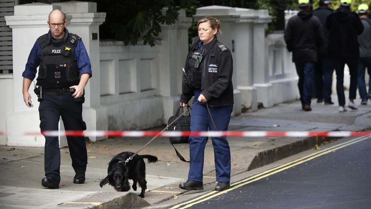 Ein Polizist und eine Polizistin gehen hinter einem rot-weißen Absperrband den Bürgersteig entlang. Die Polizistin hat den schwarzen Hund an der Leine, der den Bürgersteig beschnüffelt.