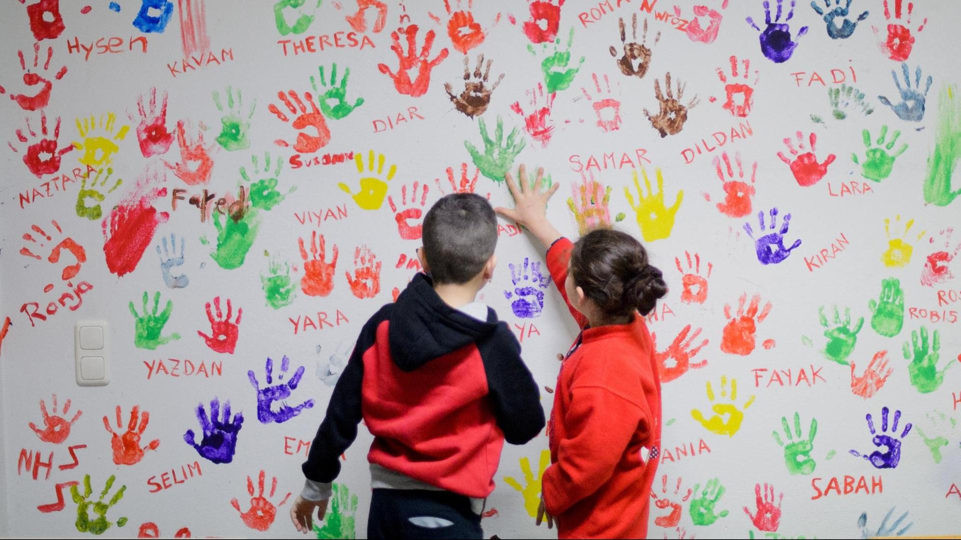 Zwei Flüchtlingskinder in einer Erstaufnahmeeinrichtung für Flüchtlinge in Hameln vor einer Wand, auf der sich junge Flüchtlinge aus der ganzen Welt mit bunten Handabdrücken verewigt haben.