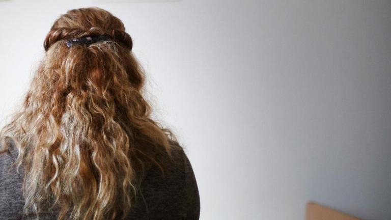 Eine Frau mit langen blonden gelockten Haaren sitzt mit dem Rücken zum Betrachter in einem Zimmer und schaut auf eine Wand mit drei Bildern