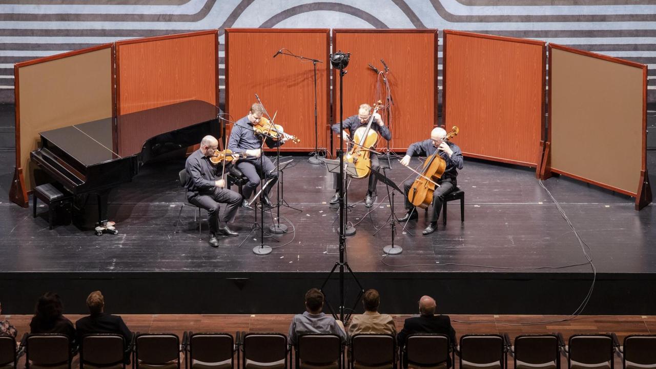Auf einer Bühne, die mit orangefarbenen Wänden im Halbkreis betont wird, spielen vier Musiker ihre Streichinstrumente.