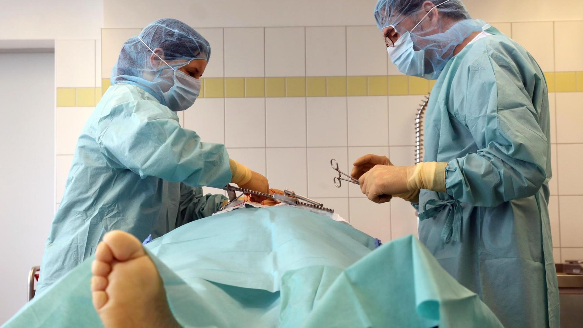 Das Foto zeigt einen Operations-Saal. In der Mitte liegt ein Mensch auf einem Tisch. Rechts und links stehen zwei Ärzte in grüner Kleidung. 