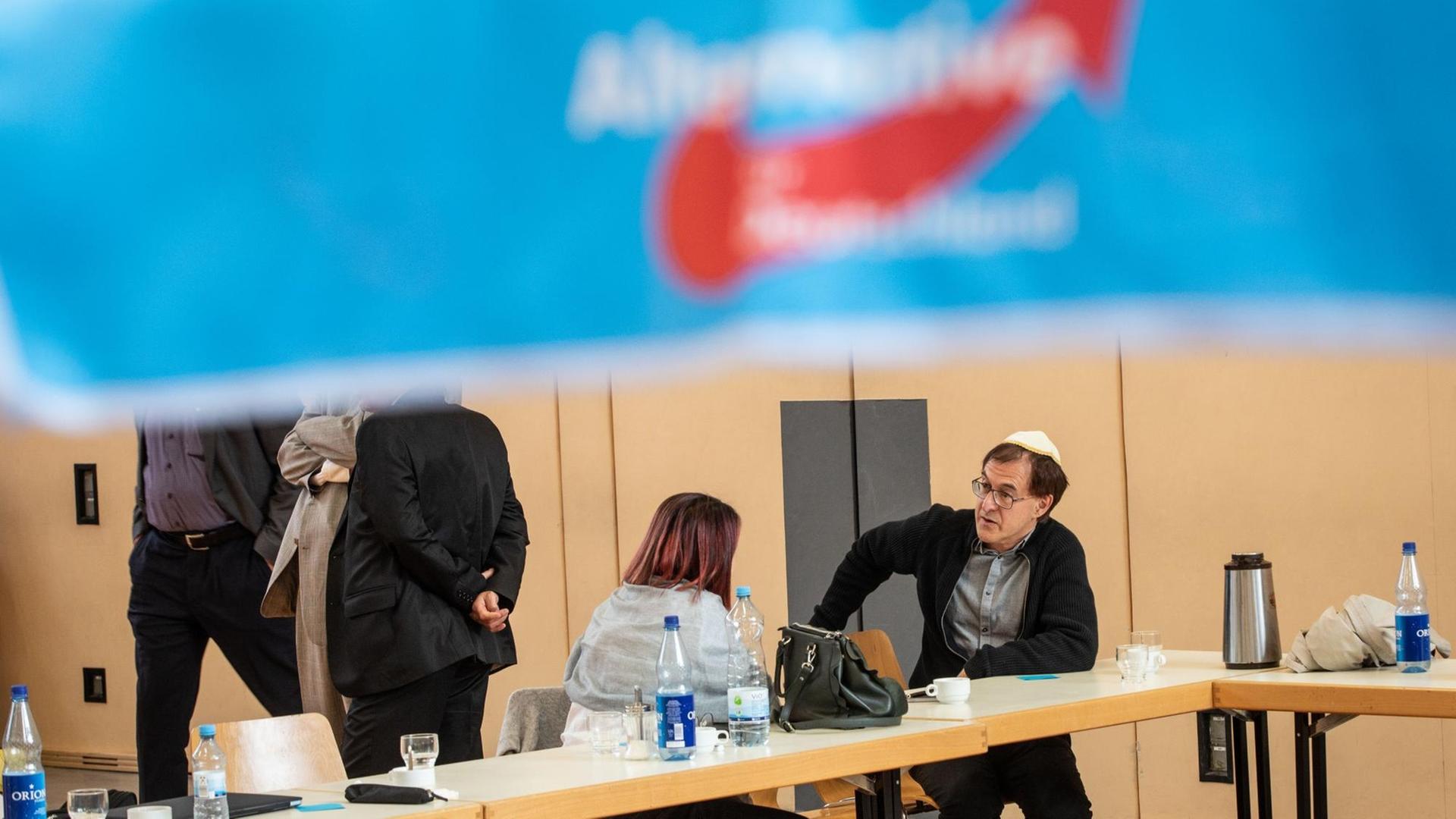 Zwei Teil-Nehmer sitzen bei der Gründungs-Veranstaltung an einem Tisch unter der einem Plakat mit dem Schrift-Zug der AfD.