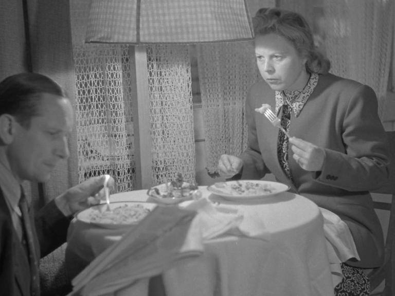 Mann und Frau sitzen gemeinsam beim Essen an einem kleinen Tisch, der Mann liest und die Frau sieht ihn leicht verärgert an.