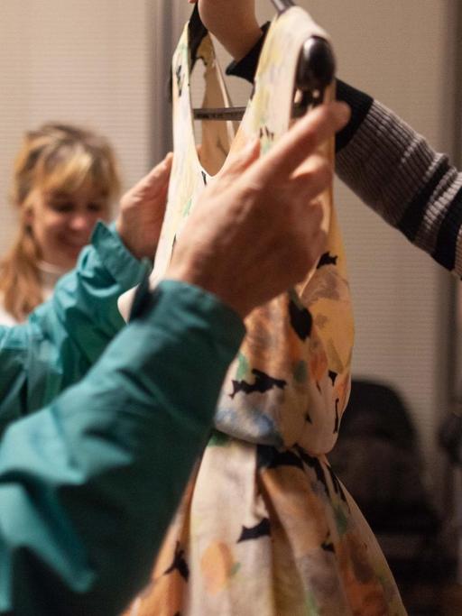 Eine Frau mit dem Blindensymbol tastet an einem Kleidungsstück, das von einer Mitarbeiterin des Berliner Ensembles hochgehalten wird.