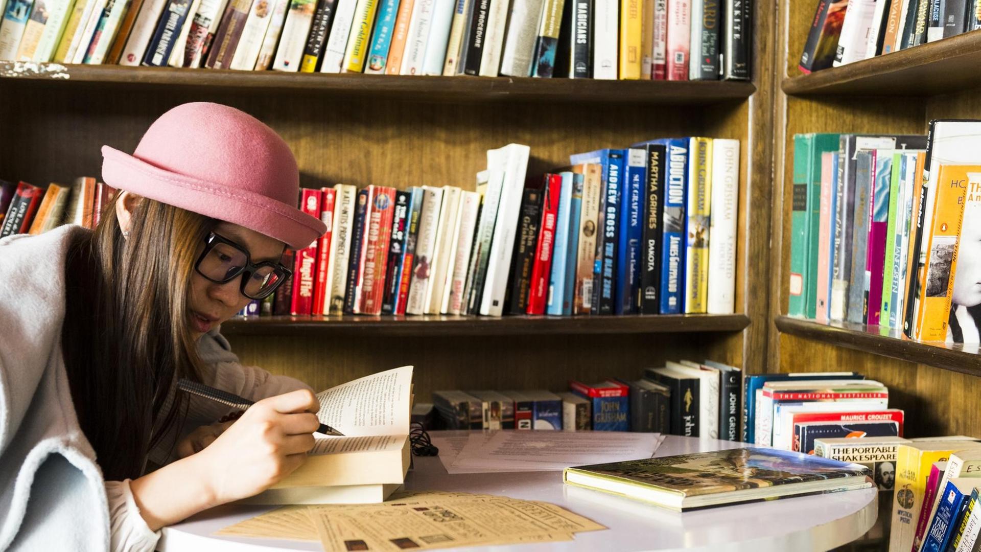Ein junge Frau, mit einem aufgeschlagenem Buch in den Händen, sitzt am Tisch vor einem vollen Bücherregal.