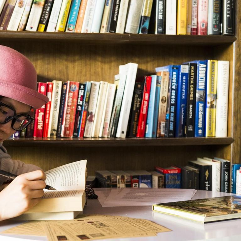 Ein junge Frau, mit einem aufgeschlagenem Buch in den Händen, sitzt am Tisch vor einem vollen Bücherregal.
