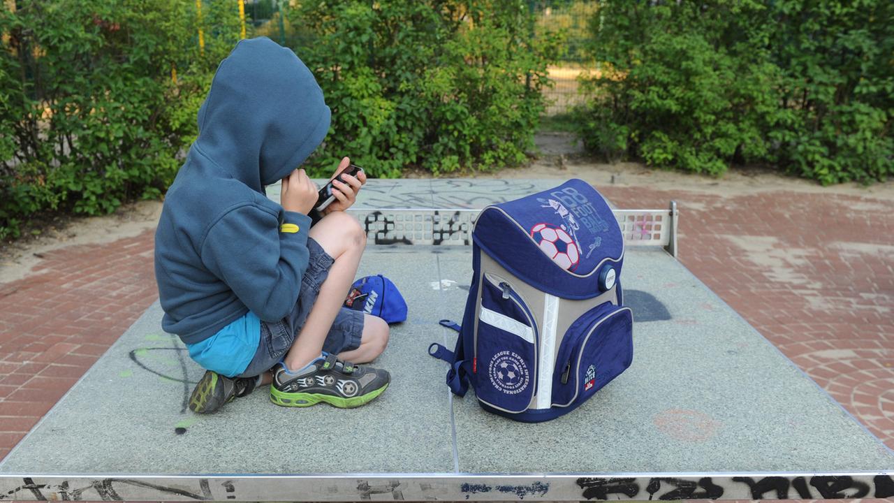 Ein Junge sitzt mit seinem Schulranzen auf einer Tischtennisplatte auf einem Spielplatz in Berlin und spielt auf einem Smartphone