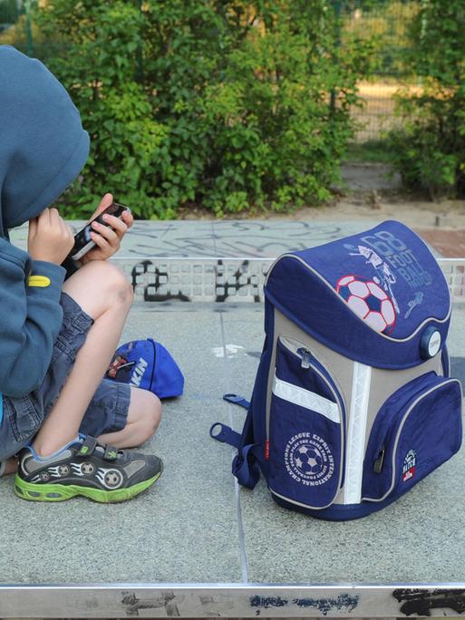 Ein Junge sitzt mit seinem Schulranzen auf einer Tischtennisplatte auf einem Spielplatz in Berlin und spielt auf einem Smartphone