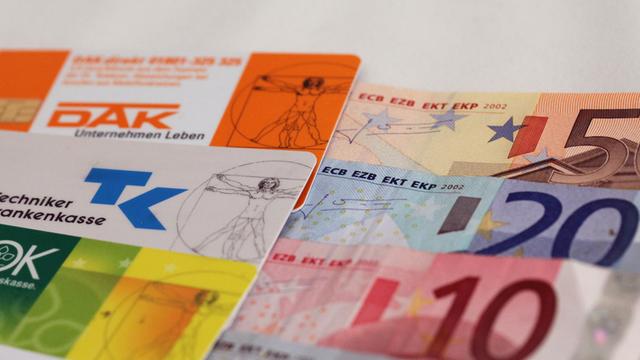 Neben Versichertenkarten verschiedener Krankenkassen liegen mehrere Geldscheine.