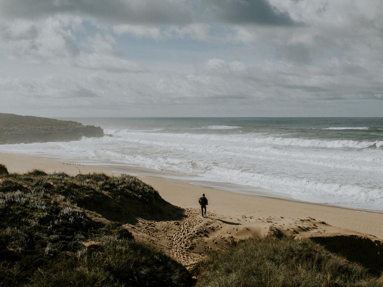 Ein Strand ist zu sehen, mit Felsen, Sand und bewölktem Himmel. Ein Mann von hinten geht in Richtung Wasser.