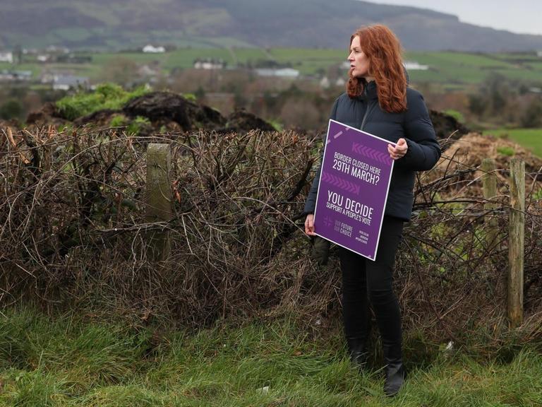 Eine Frau steht mitten in der Landschaft und protestiert mit einem Plakat an der irischen Grenze gegen den Brexit.