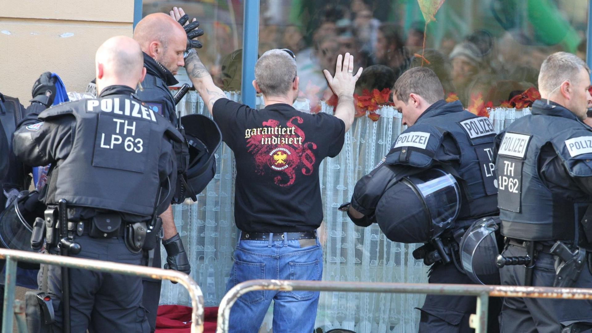 Der Besucher steht an einer großen Fensterscheibe mit den Händen nach oben. Auf dem Rücken seines Shirts steht "garantiert indiziert".Zwei Polizisten kontrollieren ihn.