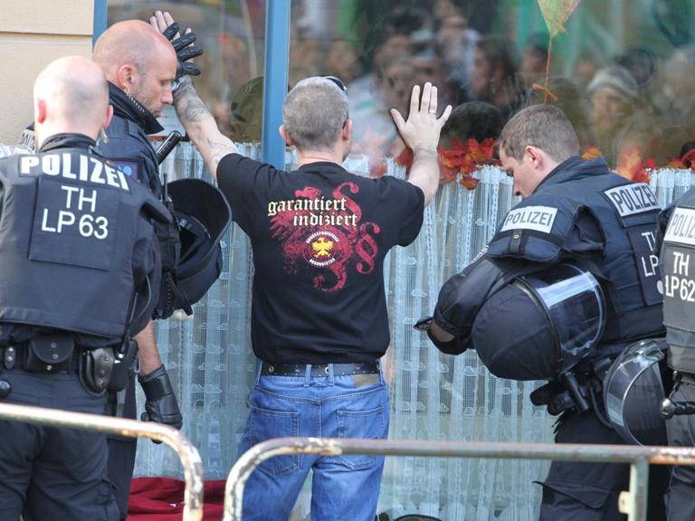 Der Besucher steht an einer großen Fensterscheibe mit den Händen nach oben. Auf dem Rücken seines Shirts steht "garantiert indiziert".Zwei Polizisten kontrollieren ihn.