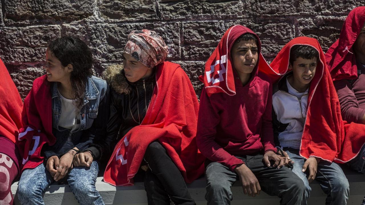 29.06.2018, Spanien, Tarifa: Junge Migranten aus Nordafrika sitzen nach ihrer Rettung in der Straße von Gibraltar im Hafen von Tarifa in Decken gehüllt an einer Mauer. Ihre Hände wurden zuvor zusammengebunden. Die EU-Staaten haben sich bei ihrem Gipfel in Brüssel darauf geeinigt, in der EU geschlossene Aufnahmelager für gerettete Bootsflüchtlinge einzurichten. Foto: Javier Fergo/dpa | Verwendung weltweit
