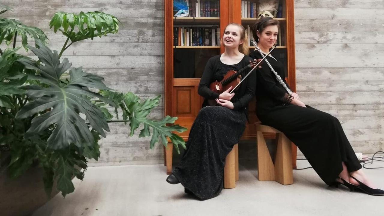 Zwei Frauen in schwarzem Kleid sitzen mit Violine und Querflöte vor einem Bücherschrank.