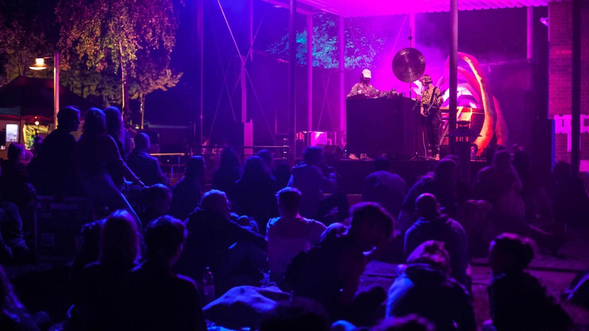 Zuschauer sitzen vor einer in lila und blau angestrahlten Bühne, auf der ein DJ und ein Gitarrist musizieren