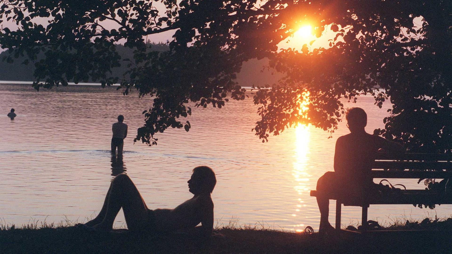 Am Stechlinsee geht die Sonne unter, ein Mensch sitzt am Ufer auf der Bank, einer liegt auf dem Rasen, einer steht im Wasser