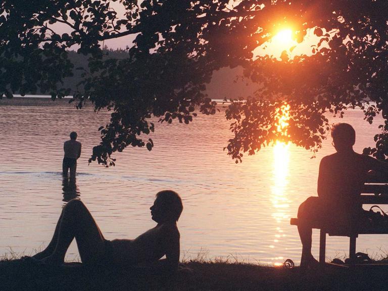 Am Stechlinsee geht die Sonne unter, ein Mensch sitzt am Ufer auf der Bank, einer liegt auf dem Rasen, einer steht im Wasser