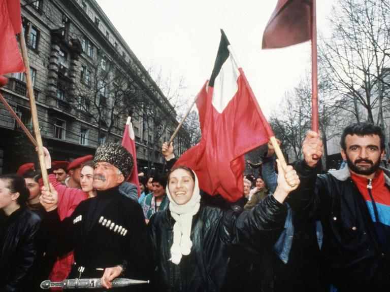 Ein Farbfoto zeigt eine Demonstration Fahnen schwenkender Menschen. in einem historischen Straßenzug. In erster Reihe ein Mann mit traditioneller Fellmütze neben einer gelöst lachenden Frau und einem bärtigen Mann