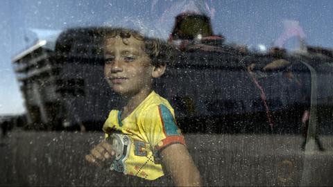 Ein Kind aus Syrien schaut in einen Bus, als gerade die Fähre 'Eleftherios Venizelos' in dessen Scheibe gespiegelt wird.