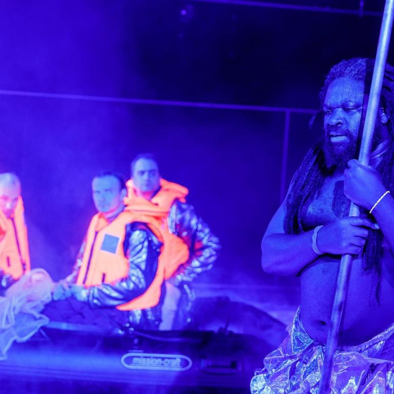 Eine Szene aus Konstantin Küsperts neuem Stück "Sklaven leben" am Schauspiel Frankfurt, Januar 2019. Vier europäisch aussehende Menschen sitzen in Schwimmwesten in einem Schlauchboot. Im Vordergrund steht ein Schwarzer mit Rastalocken und hält einen Stab in den Händen.