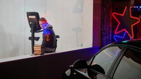 Eine Schauspielerin sitzt auf einen Fenstersims vor einem geparkten Auto. neben ihr liegt ein Bücherstapel. An der Hauswand leuchtet ein großer roter Stern.