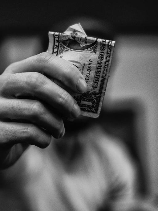 Eine Hand hält einen Dollarschein in die Kamera, der Kopf versteckt sich dahinter.