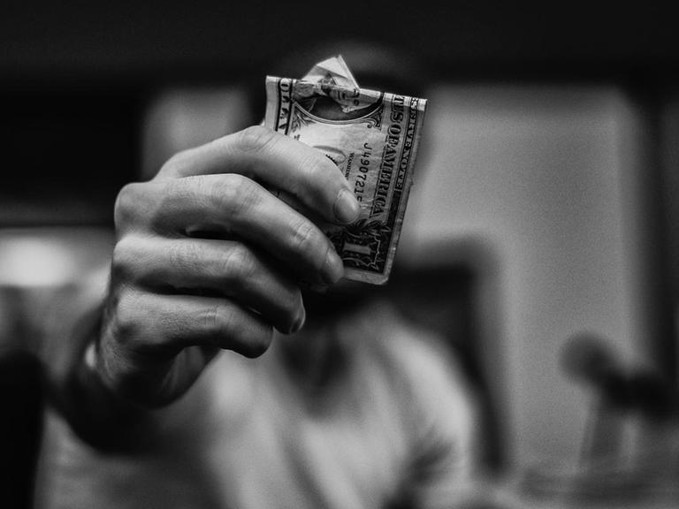 Eine Hand hält einen Dollarschein in die Kamera, der Kopf versteckt sich dahinter.