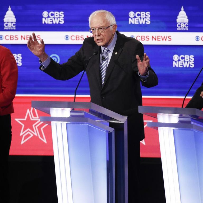Elizabeth Warren Bernie Sanders und Joe Biden stehen an Redepulten nebeneinander. Sanders hat beide Arme vor sich ausgebreitet.