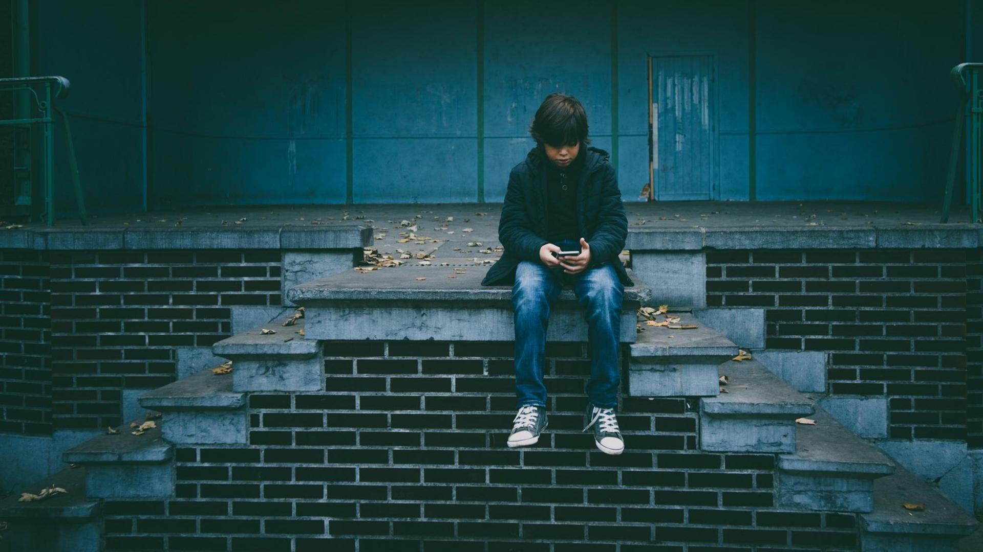 Ein Junge sitzt auf einer Treppe und schaut auf sein Smartphone.
