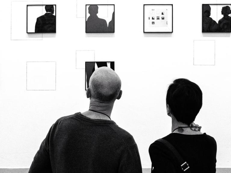 Das schwarz-weiße Bild zeigt zwei Personen, die auf eine Wand mit vielen ungeordneten Bilderrahmen gucken.
