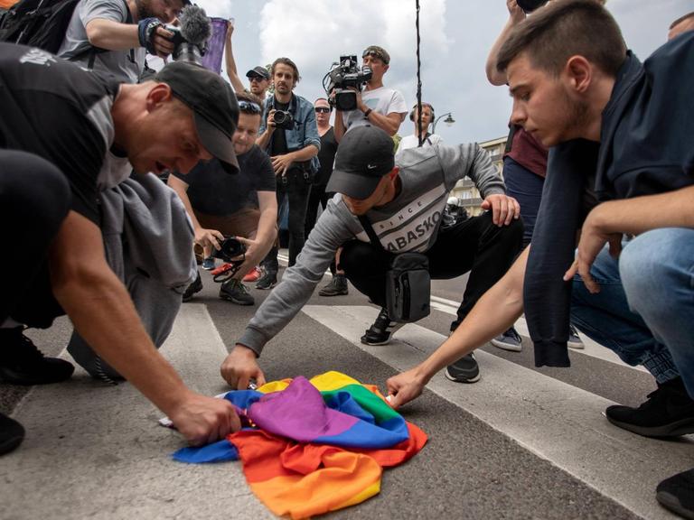 Eine Gruppe junger Männer hält Feuerzeuge an eine Regenbogenflagge, die zusammengeknüllt auf einem Zebrastreifen liegt.