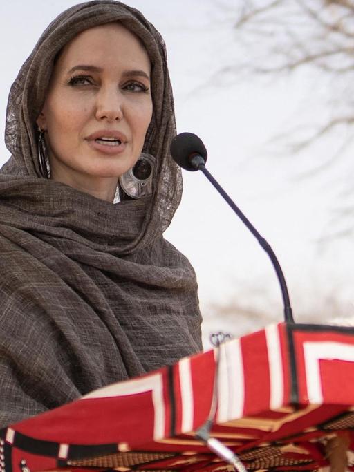 Angelina Jolie seht mit einem Kopftuch bekleidet vor einem Pult mit einem Mikrofon.