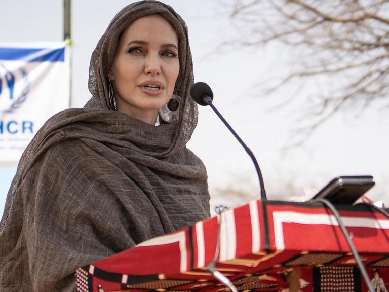 Angelina Jolie seht mit einem Kopftuch bekleidet vor einem Pult mit einem Mikrofon.