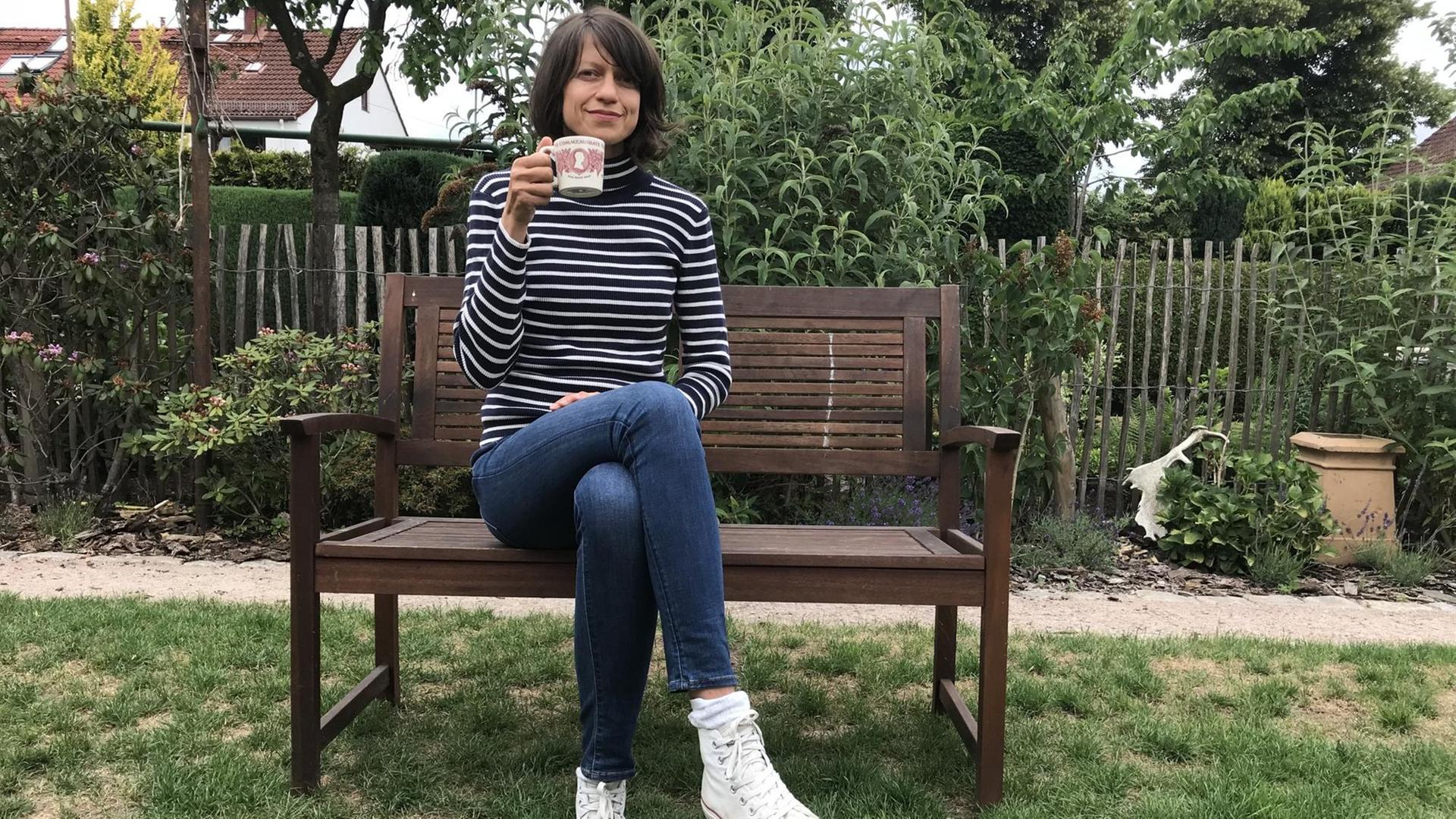 Dlf-Autorin Louise Brown sitzt auf einer Bank und mit Teetasse in der Hand in einem Garten