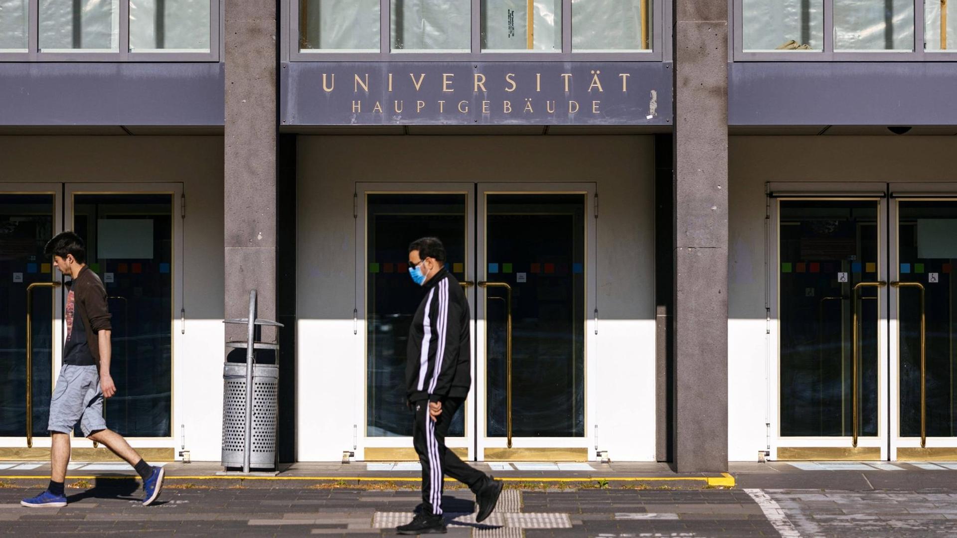 Der fast menschenleere Eingang der Albertus Magnus Universität zu Köln. Nur zwei junge Männer, davon einer mit Mundschutz, laufen an dem Gebäude vorbei.