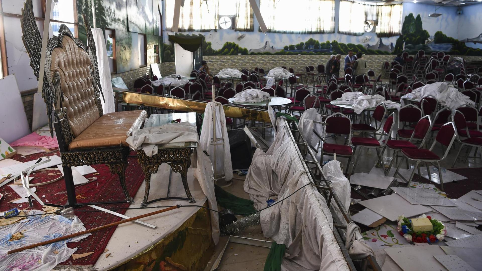 Im Vordergrund des Bildes ist ein durch eine Explosion zerstörter Hochzeitssaal in Kabul zu sehen, im Hintergrund untersuchen Männer den Raum.