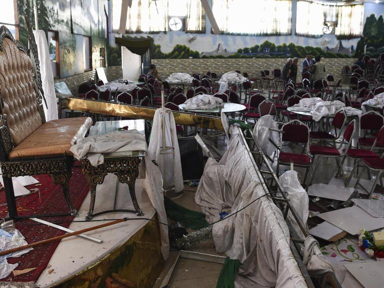 Im Vordergrund des Bildes ist ein durch eine Explosion zerstörter Hochzeitssaal in Kabul zu sehen, im Hintergrund untersuchen Männer den Raum.