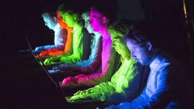 Bunt angestrahlte Musiker sitzen konzentriert vor Laptops.