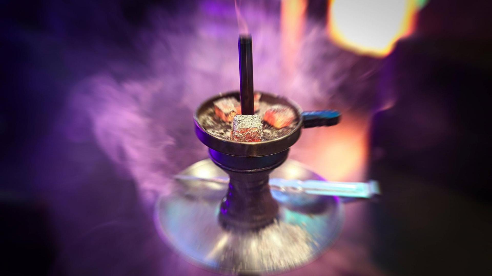 Eine Shisha Wasserpfeife mit drei glühenden Kohlen auf dem Kaminaufsatz, Rauch und Neonlichter im Hintergrund.