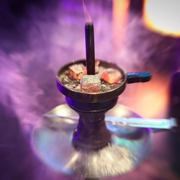 Eine Shisha Wasserpfeife mit drei glühenden Kohlen auf dem Kaminaufsatz, Rauch und Neonlichter im Hintergrund.