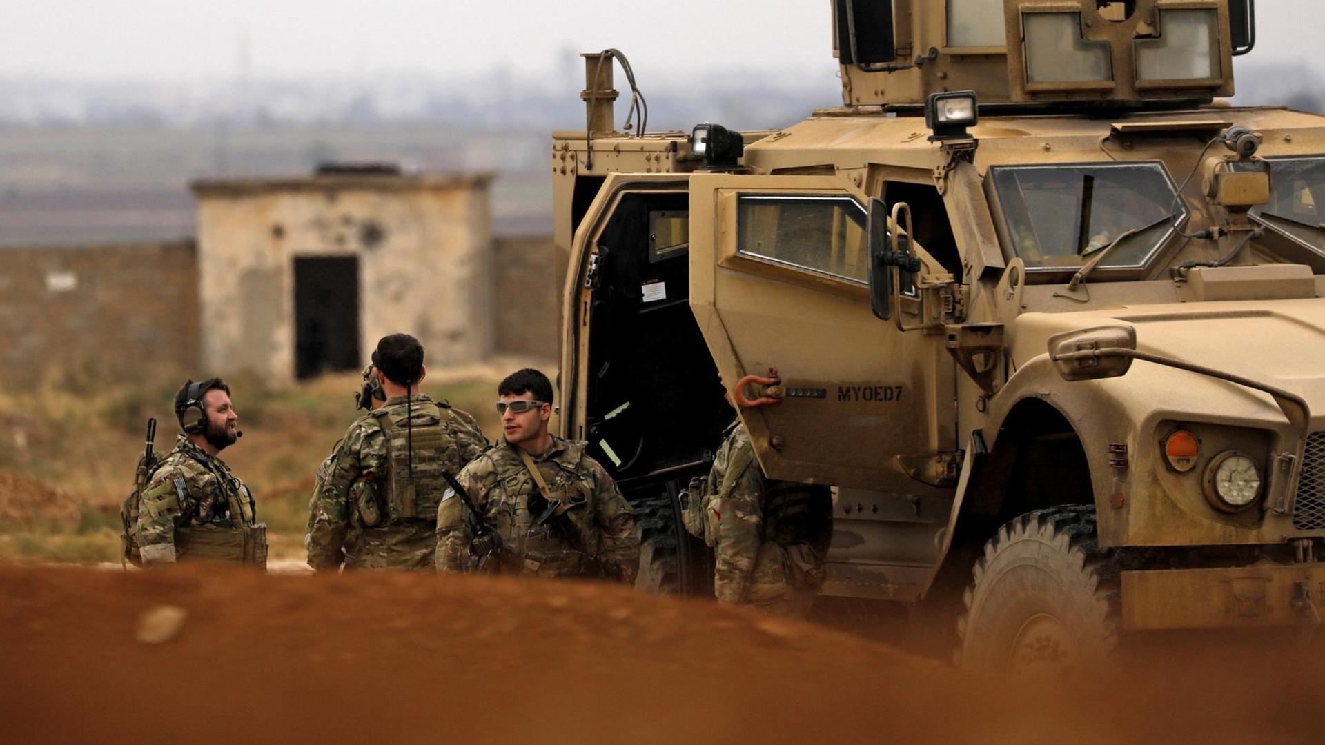 Drei Kämpfer in Tarnuniformen stehen vor einem sandfarbenen gepanzerten Fahrzeug vor einer kleinen Hütte. 