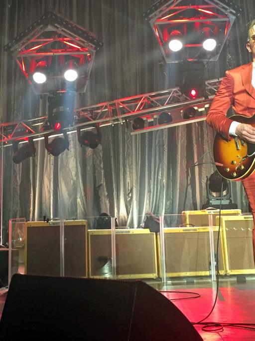 Ein Mann im roten Anzug steht an auf einem Bühne einem Mikrofon, er singt und spielt Gitarre. Im Hintergrund steht ein Schlagzeug und eine ganze Reihe Gitarrenverstärker.