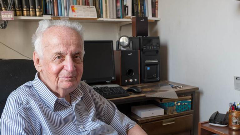 Ein älterer Mann sitzt in einem Arbeitsstuhl. Hinter ihm ist ein Computer und ein Bücherregel. Es ist Ivan Ivanji in seinem Arbeitszimmer in Belgrad.