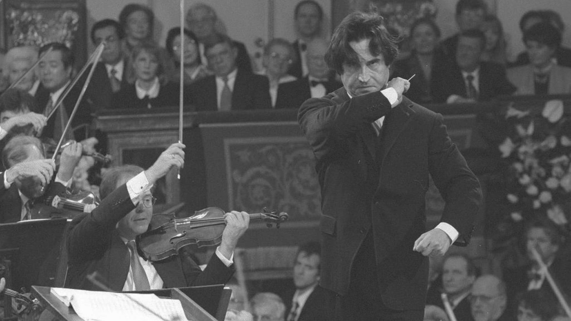 Schwarz-weiß-Aufnahme mit dem Dirigenten rechts im Bild, links sitzen die 1. Geiger mit erhobenen Bögen, im Hintergrund sitzt das Publikum. Der Dirigent trägt einen schwarzen Anzug, ein weißes Hemd und Schlips, die dunklen Haare fallen ihm ins Gesicht, der Taktstock ist erhoben zum Schlussakkord.
