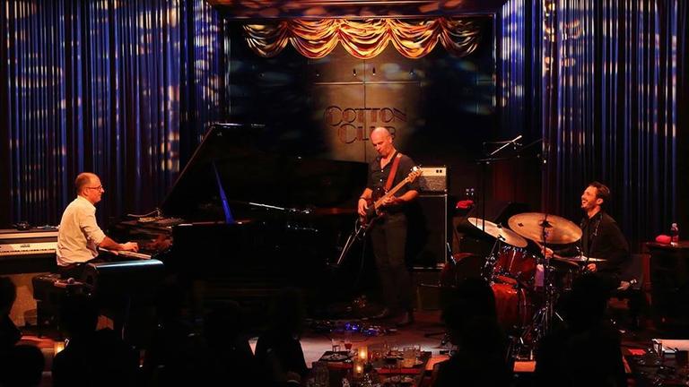 Das Trio steht mit Flügel, E-Gitarre und Schlagzeug auf einer gelb-blau beleuchteten Bühne.