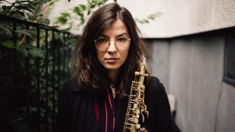 Eine junge, dunkelhaarige Frau mit Brille und einem Saxofon in der Hand steht in einem Hinterhof, im Bildhintergrund Pflanzen und eine Hauswand.