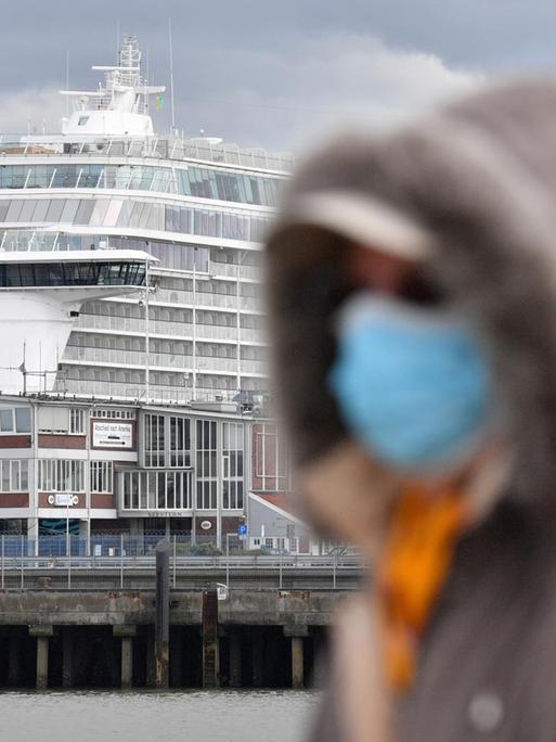Eine Person mit Atemschutzmaske geht an dem Kreuzfahrtschiff, welches am Dock liegt, vorbei.