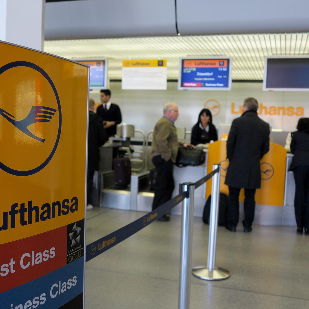 Check-in-Schalter der Lufthansa, im Vordergrund ein Lufthansa-Schild, im Hintergrund der Schalter mit Mitarbeitern und Passagieren.